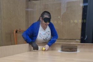 Eine Frau mit dunkler Augenbinde und Handschuhen steht hinter einem Showdown-Tisch. Vor ihr liegt ein kleiner gelber Ball, den sie mit beiden Händen berührt.